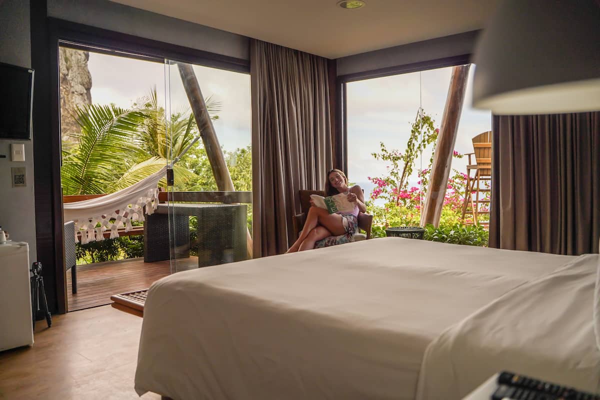 Un dormitorio con una cama grande, una hamaca y vistas a la vegetación y flores por la ventana.