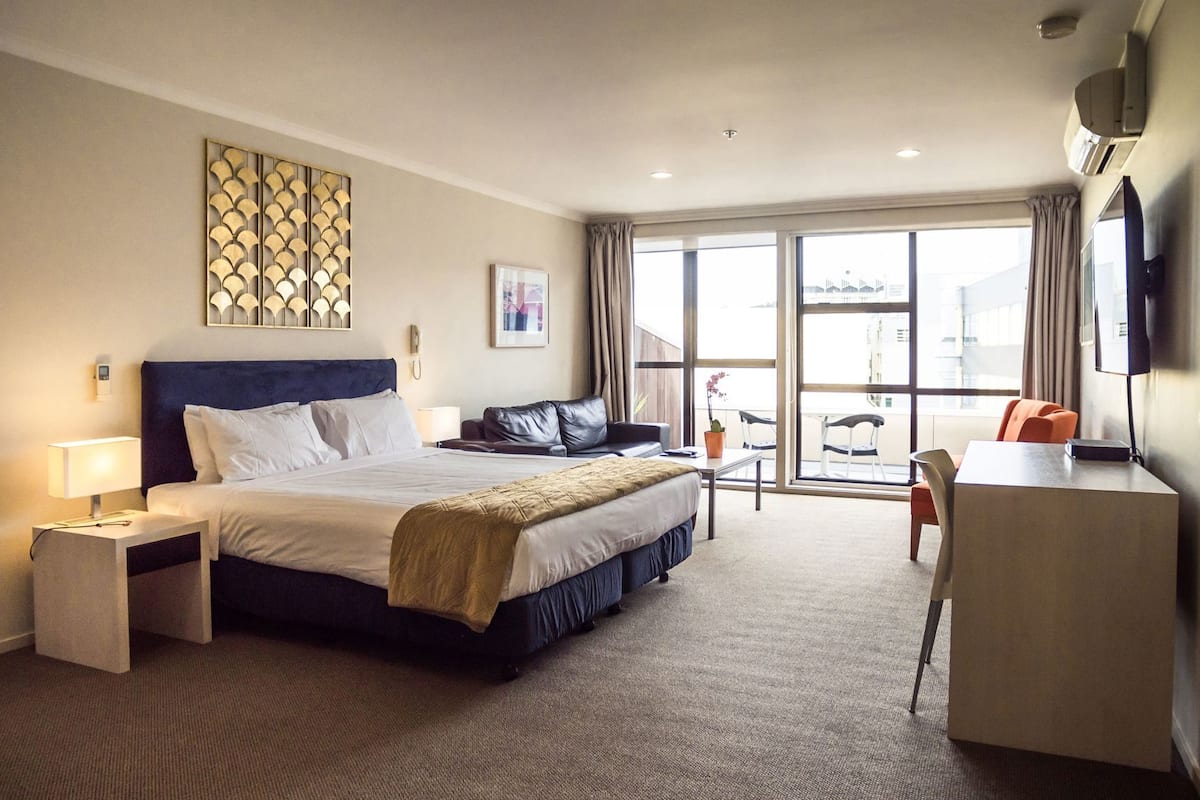 A hotel room with a bed, bedside tables, a desk, a chair, a large window, and a view of the city.