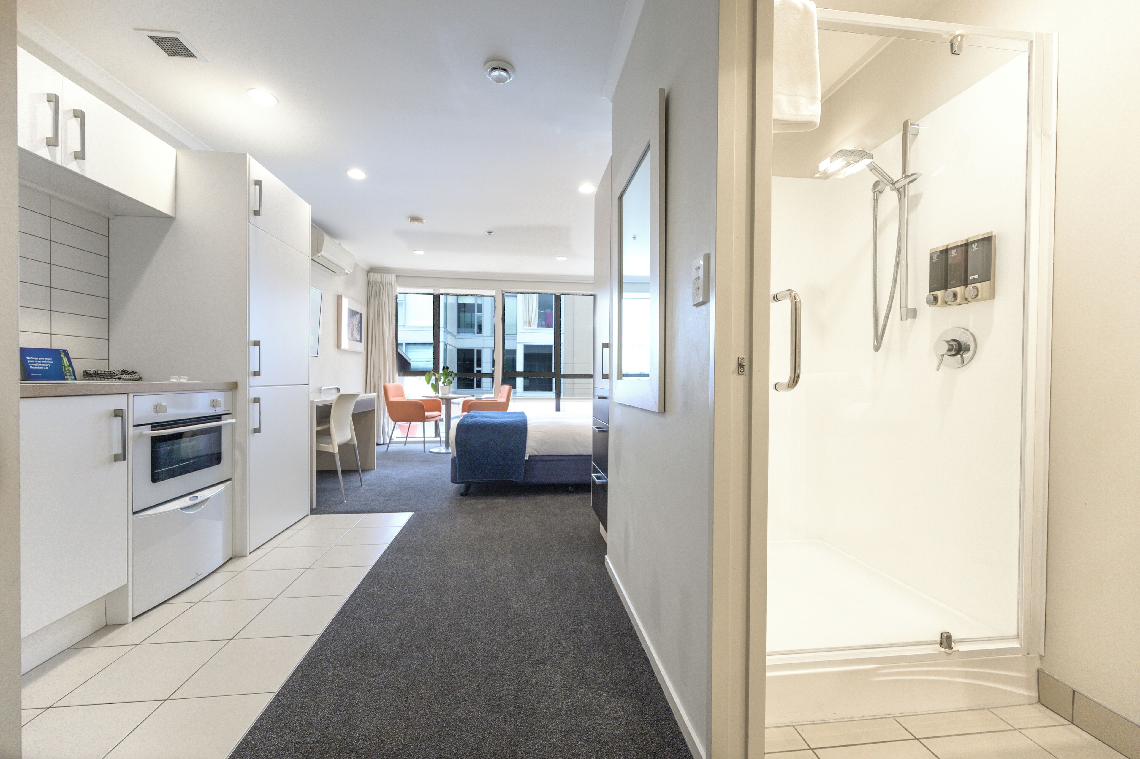 A modern hotel room with a kitchenette, a shower area, a bed, and a seating area with a view of the city.