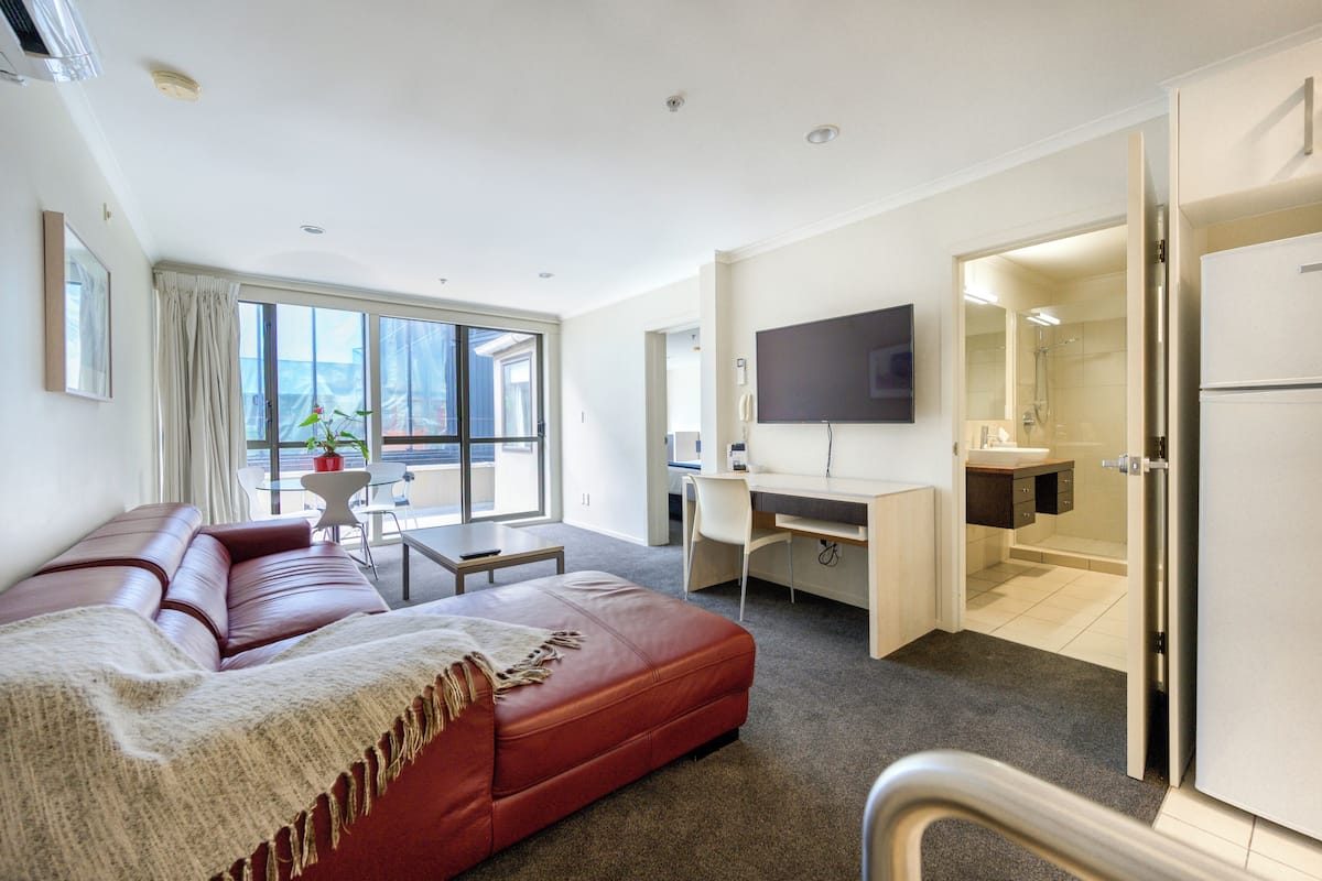 A modern hotel room with a red leather sofa, a desk with a computer, a flat-screen TV, and a bathroom visible through an open door.