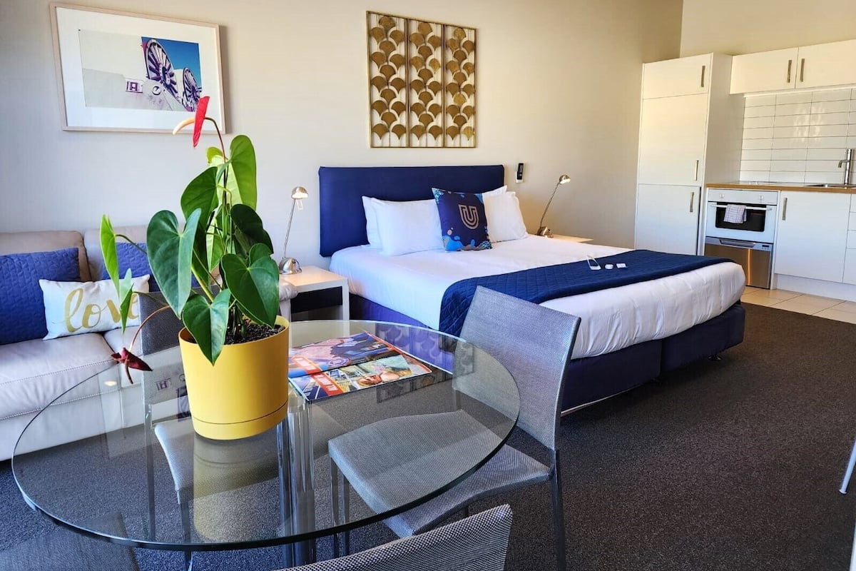 A modern hotel room with a bed, a sofa, a glass table, and a kitchen area.