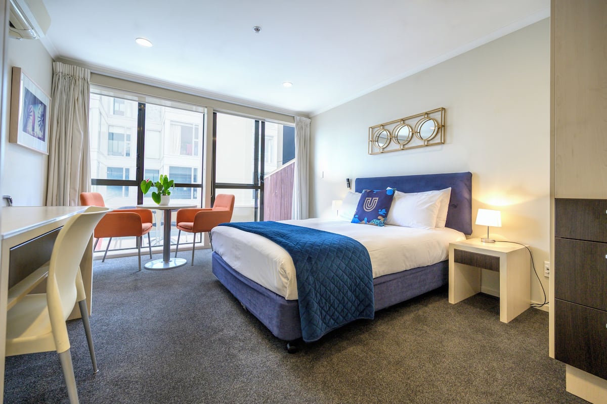 A hotel room with a bed, bedside table, lamp, and a desk with chairs.