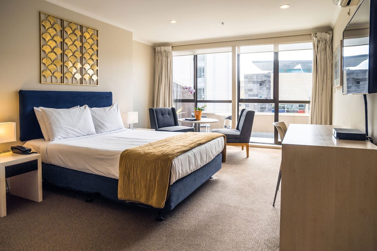 A hotel room with a large bed, a desk, a chair, a small table, and a window with a view of buildings.