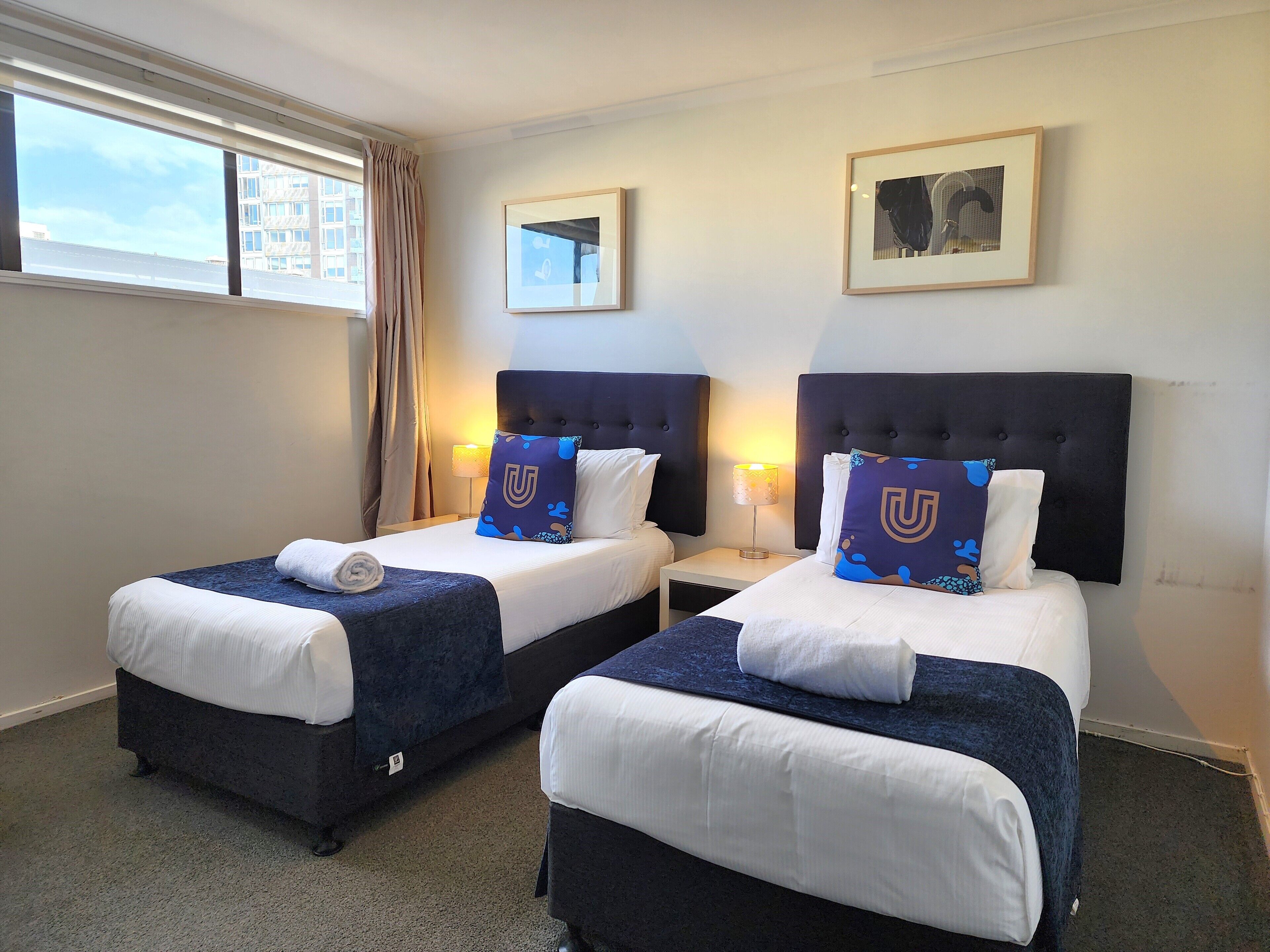 A hotel room with two beds, each with a dark headboard, white bedding, and a blue pillow with a 'U' logo. There are two bedside lamps, a framed picture on the wall, and a window with curtains.
