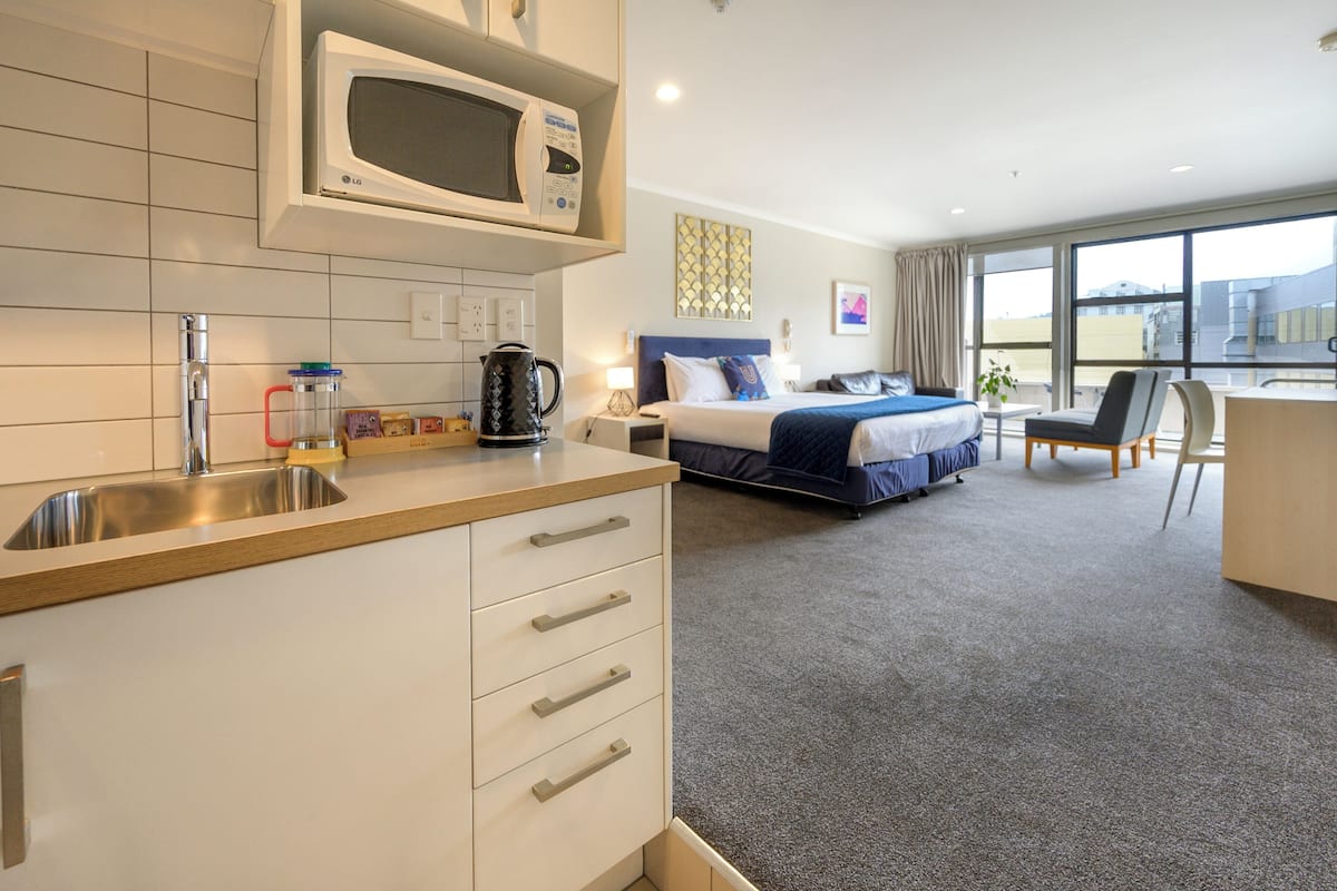 A hotel room with a kitchenette, a bed with a blue headboard, a desk with a chair, and a large window offering a view of the outside.