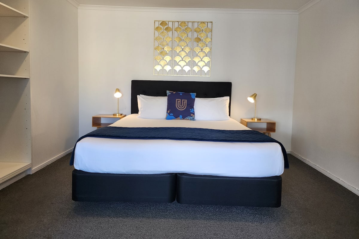A neatly arranged hotel room with a large bed, two bedside tables with lamps, a decorative wall piece, and built-in shelving.