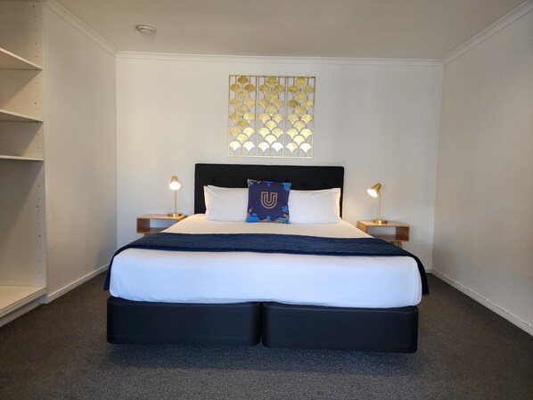 Habitación de hotel ordenada con una cama grande, dos mesitas de noche con lámparas, un adorno en la pared y estanterías empotradas.