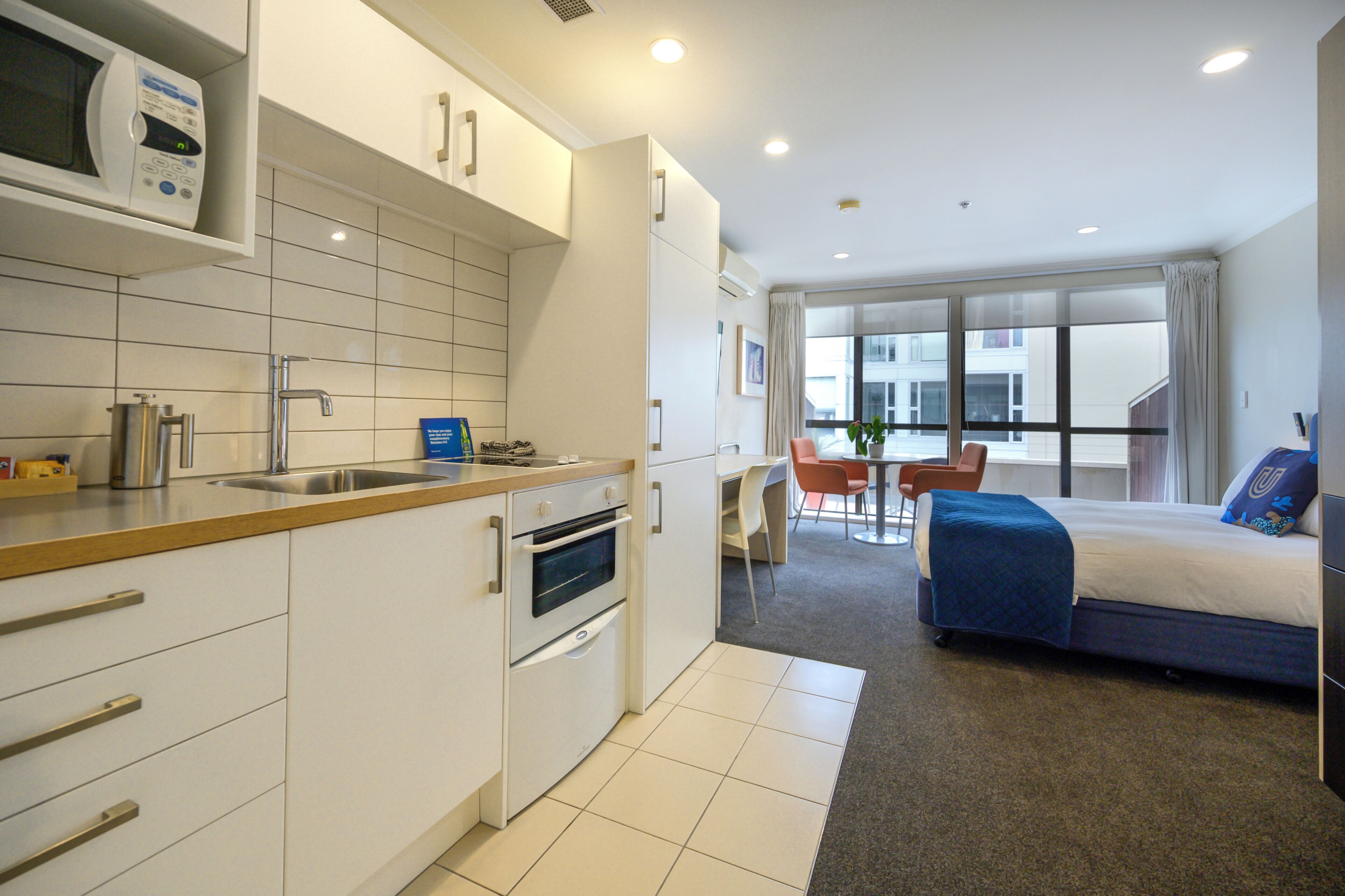 A modern hotel room with a kitchenette, a bed with a blue blanket, a dining area with a table and chairs, and large windows offering a view of the city.