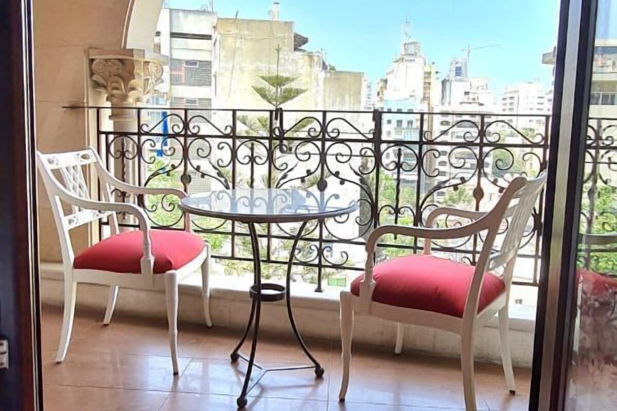 A balcony with two white chairs and a table, a red rug, and a view of buildings through a glass door.