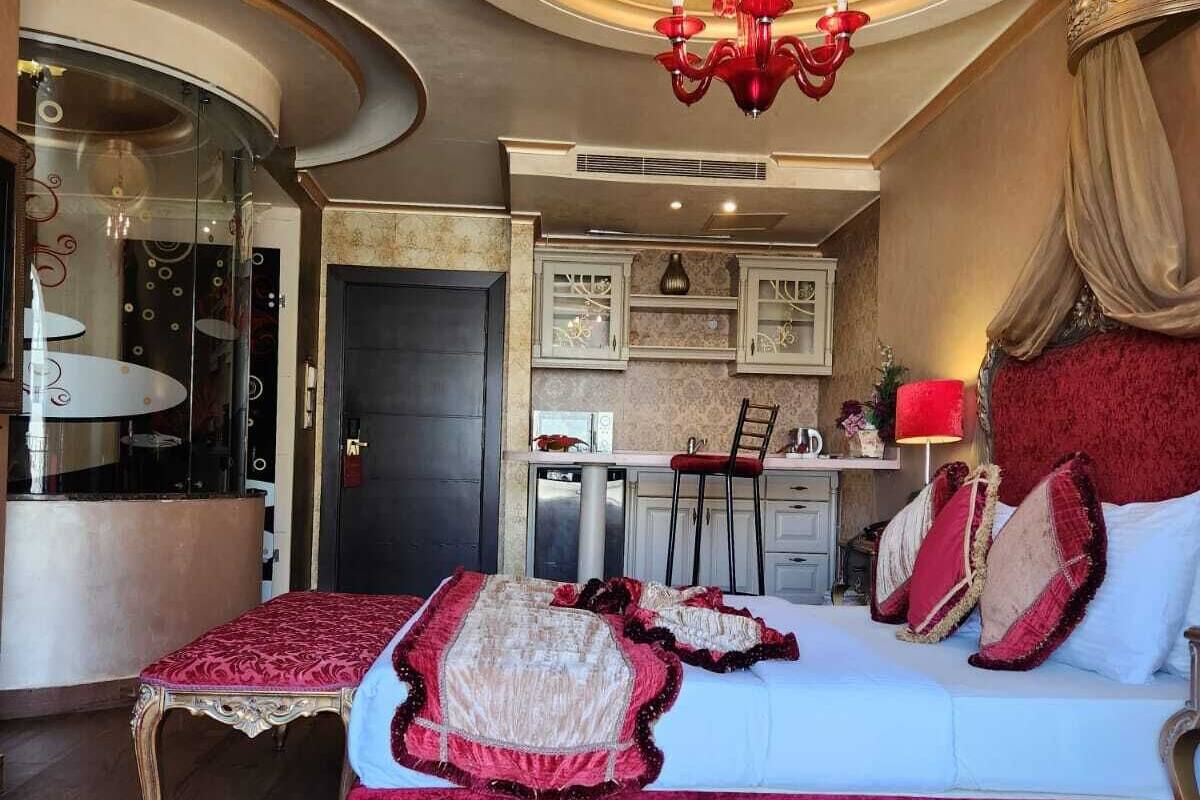 A bedroom with a large bed, a chandelier, a red carpet, and a small kitchenette.