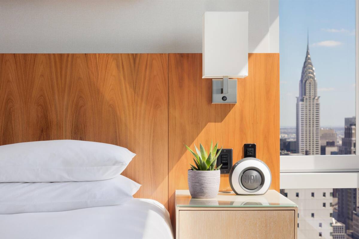 Quarto de hotel com cama, luminária na parede, planta, alto-falante e vista para a cidade.