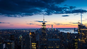 Une vue sur une ville au crépuscule, avec de hauts gratte-ciel bien visibles, dont une tour illuminée et une flèche, sous un ciel teinté des couleurs du soir.