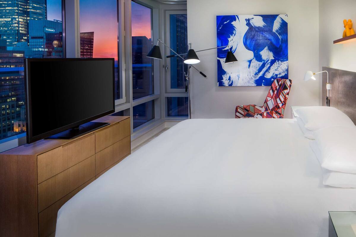 Quarto de hotel com uma cama grande, uma televisão em um móvel de madeira, vista para a cidade, um quadro e um vaso com flores.