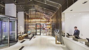 A modern hotel lobby with a curved reception desk, glass elevator, and a seating area with a coffee machine. - Hyatt Centric Times Square New York (New York)
