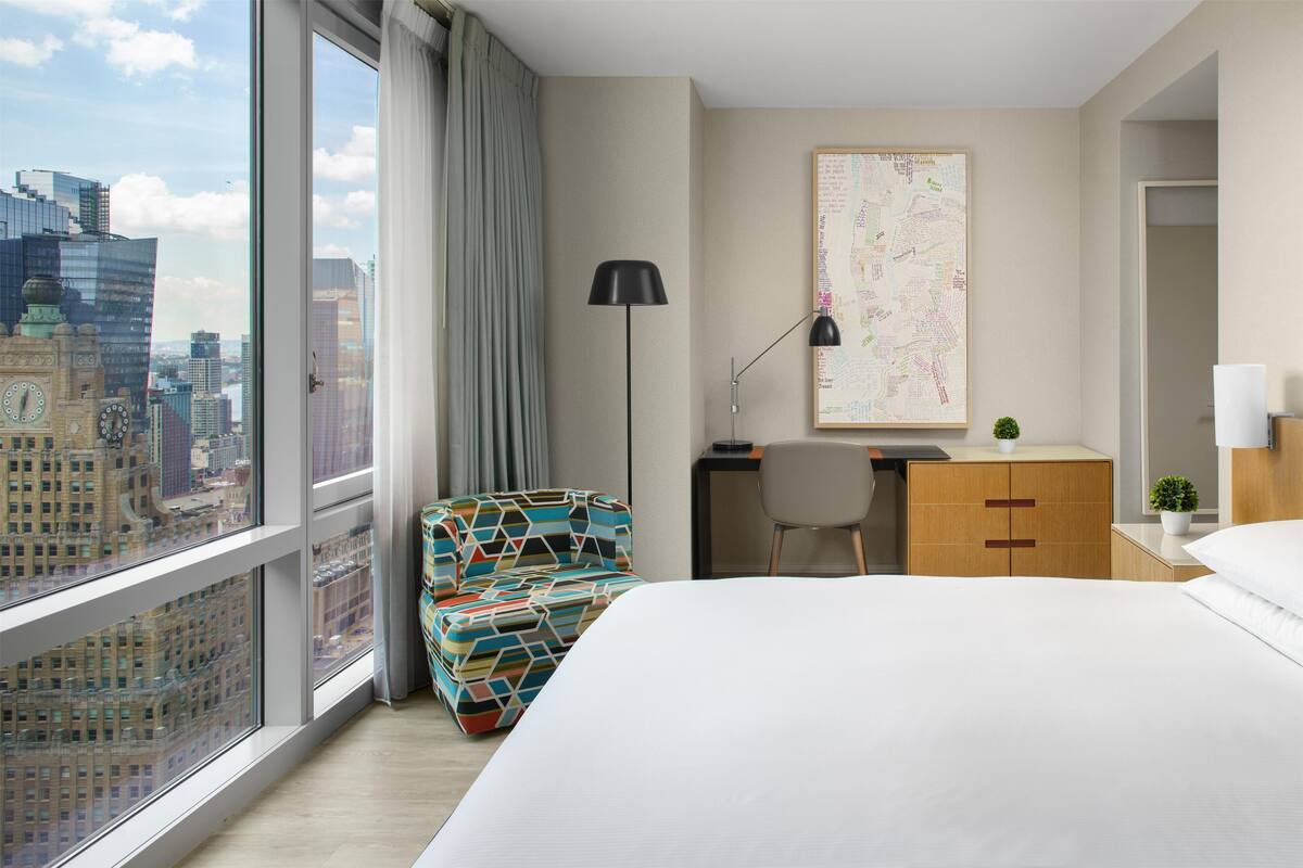Quarto de hotel com uma janela grande, uma cama, uma escrivaninha, uma cadeira e um mapa na parede.
