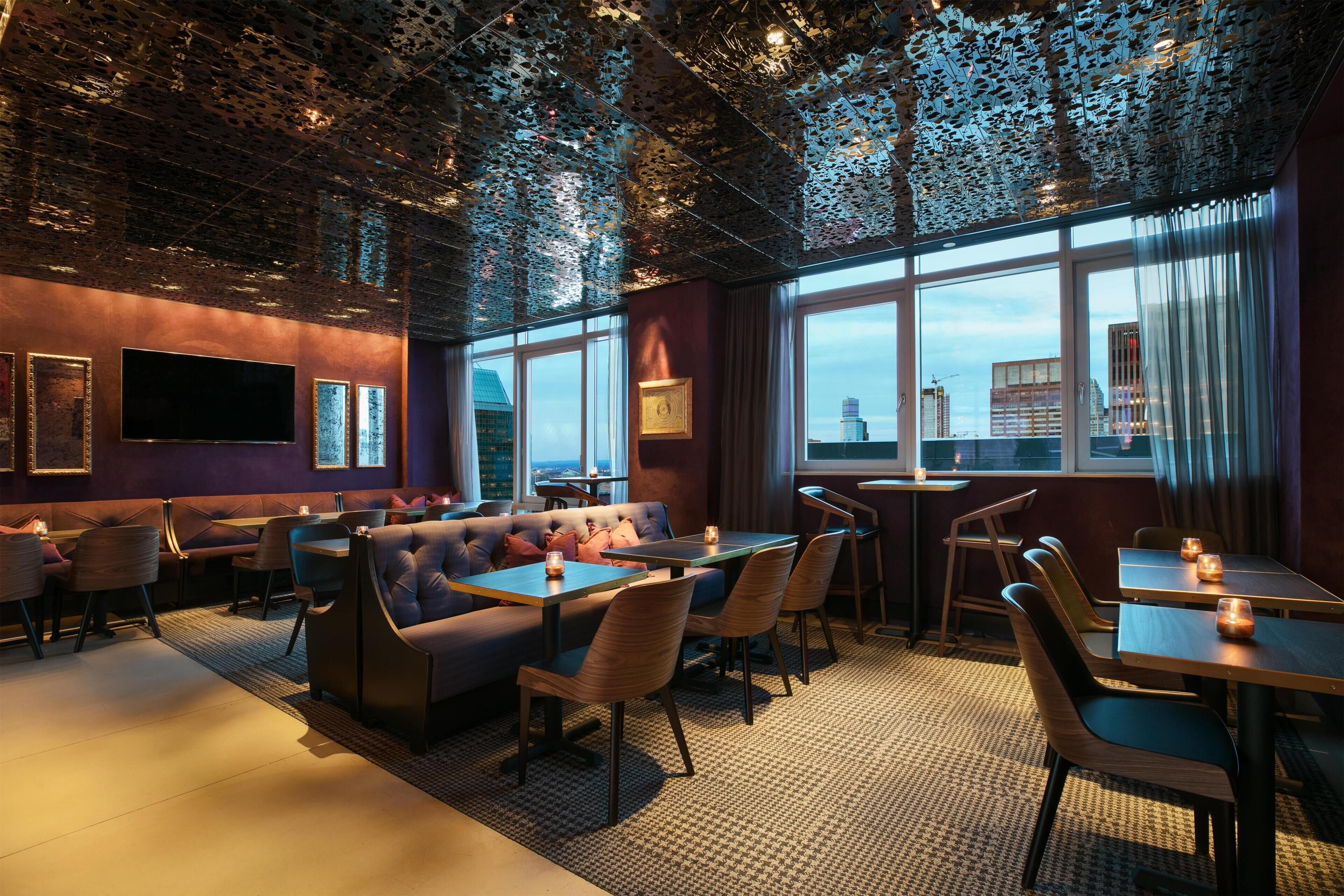 A modern restaurant with a high ceiling, large windows, and a city view.