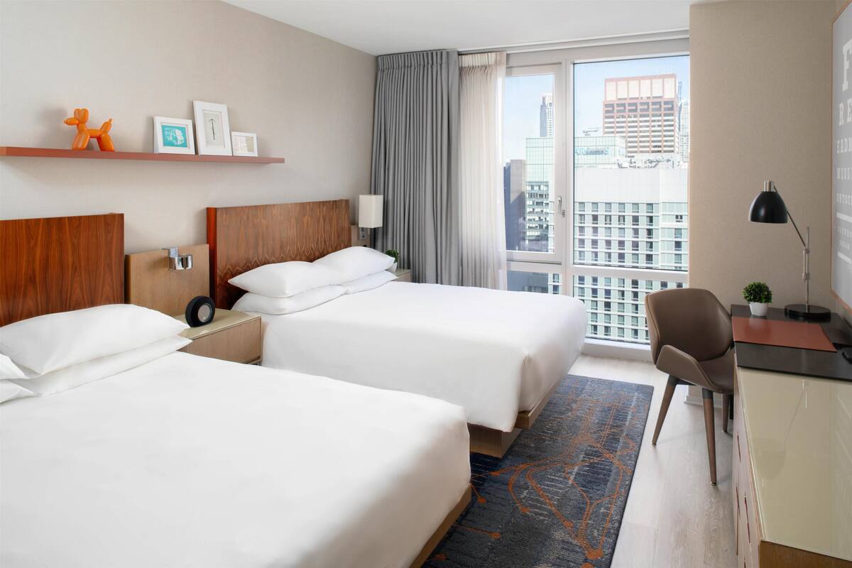 Quarto de hotel com duas camas, uma escrivaninha, uma cadeira, uma janela com vista para a cidade e cortina.