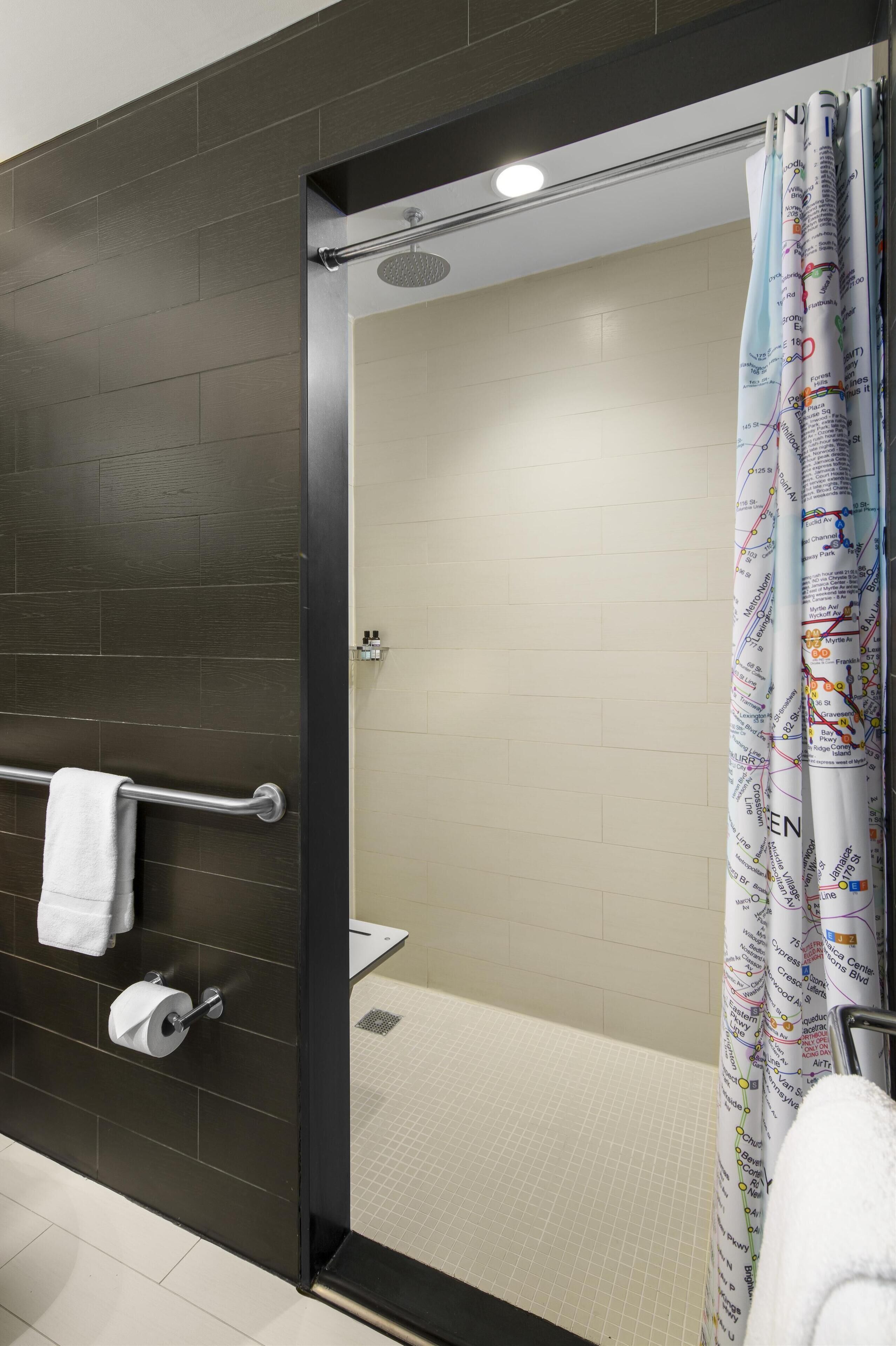 A modern bathroom with a dark tiled wall, a glass shower enclosure, a toilet paper holder, and a shower curtain with a map design.