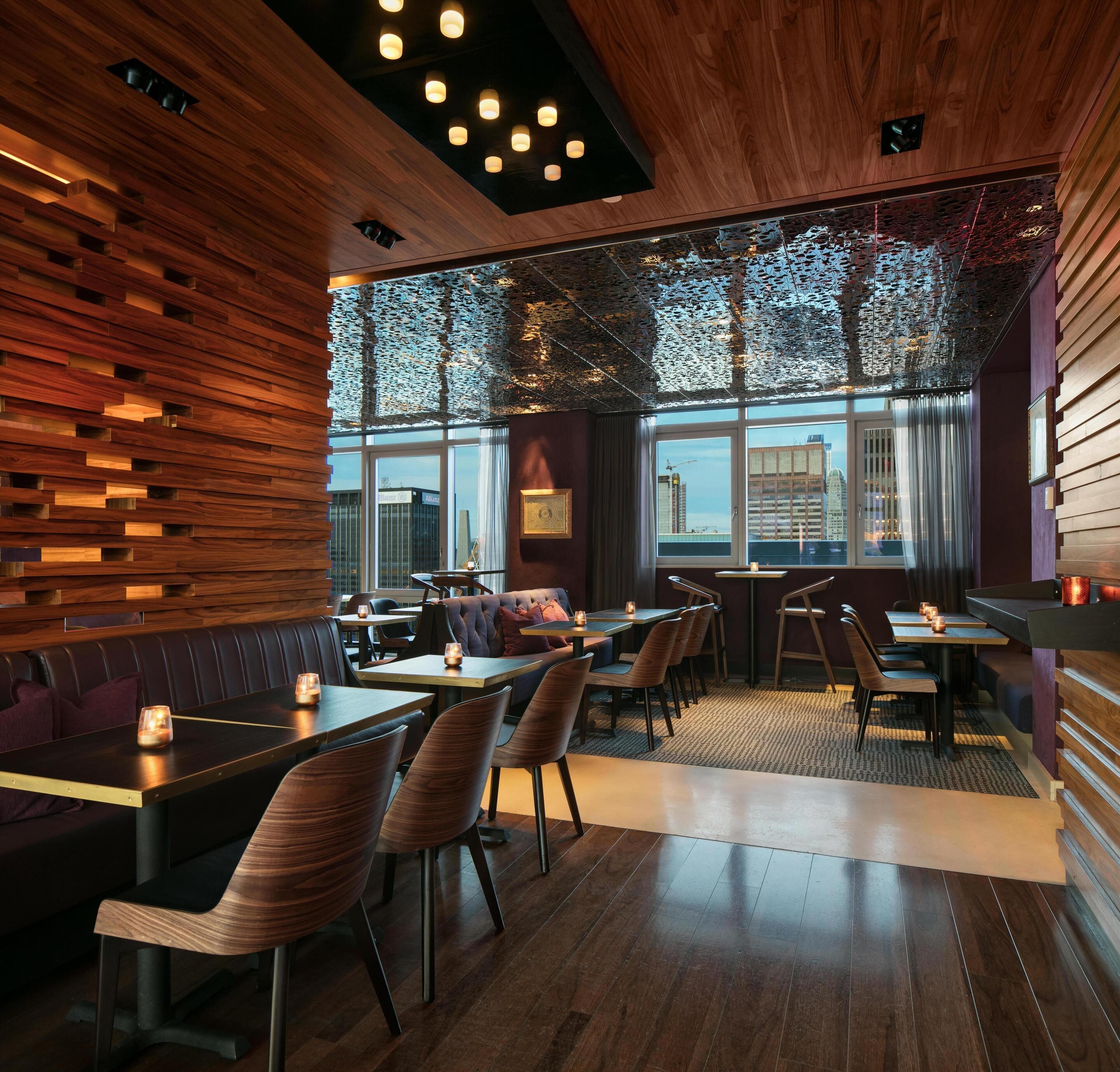 A restaurant with wooden walls, tables, and chairs, and large windows with a view of buildings.