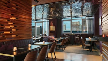 Um restaurante com paredes, mesas e cadeiras de madeira, e janelas amplas com vista para edifícios.