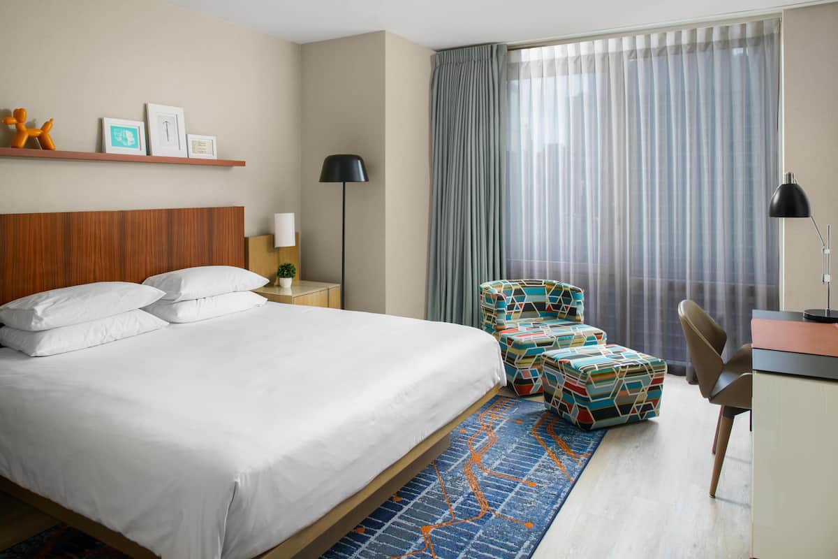 Quarto de hotel com cama, escrivaninha, cadeira, luminária, mesa lateral, prateleira na parede com quadros e janela com cortinas.