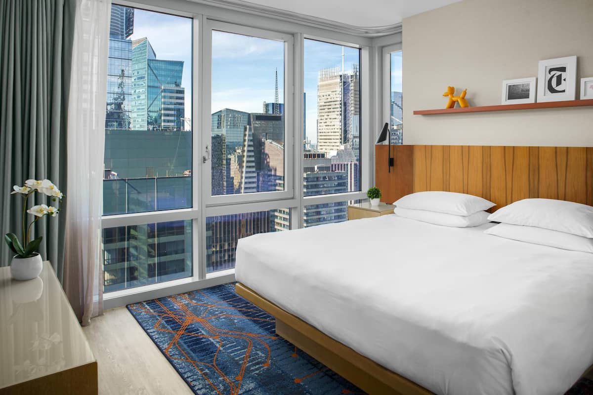 Quarto de hotel com uma cama grande, uma mesa de cabeceira, uma planta em vaso e vista para a cidade através de janelas amplas.