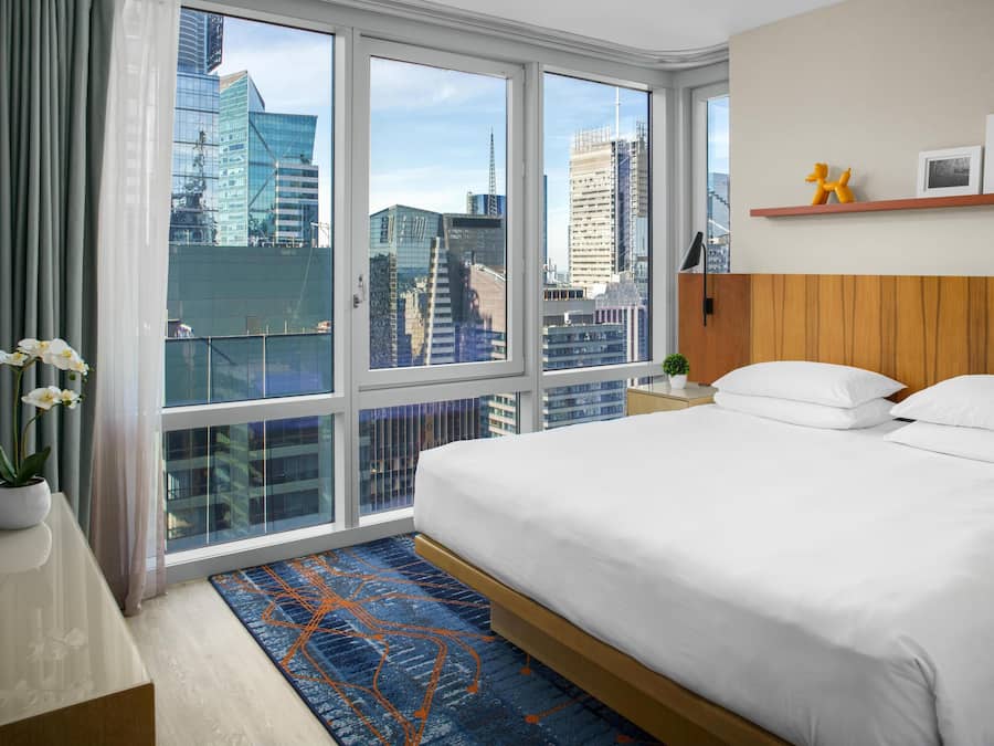 Ein Hotelzimmer mit einem großen Bett, einem Nachttisch, einer Topfpflanze und Blick auf die Stadt durch große Fenster.