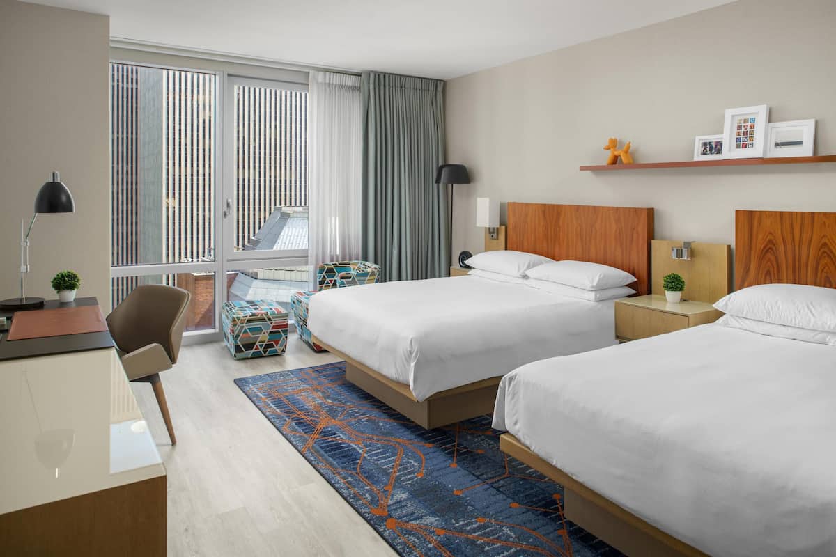 Quarto de hotel com duas camas, uma escrivaninha, uma cadeira, uma mesinha, uma luminária, uma planta e uma janela com cortinas.