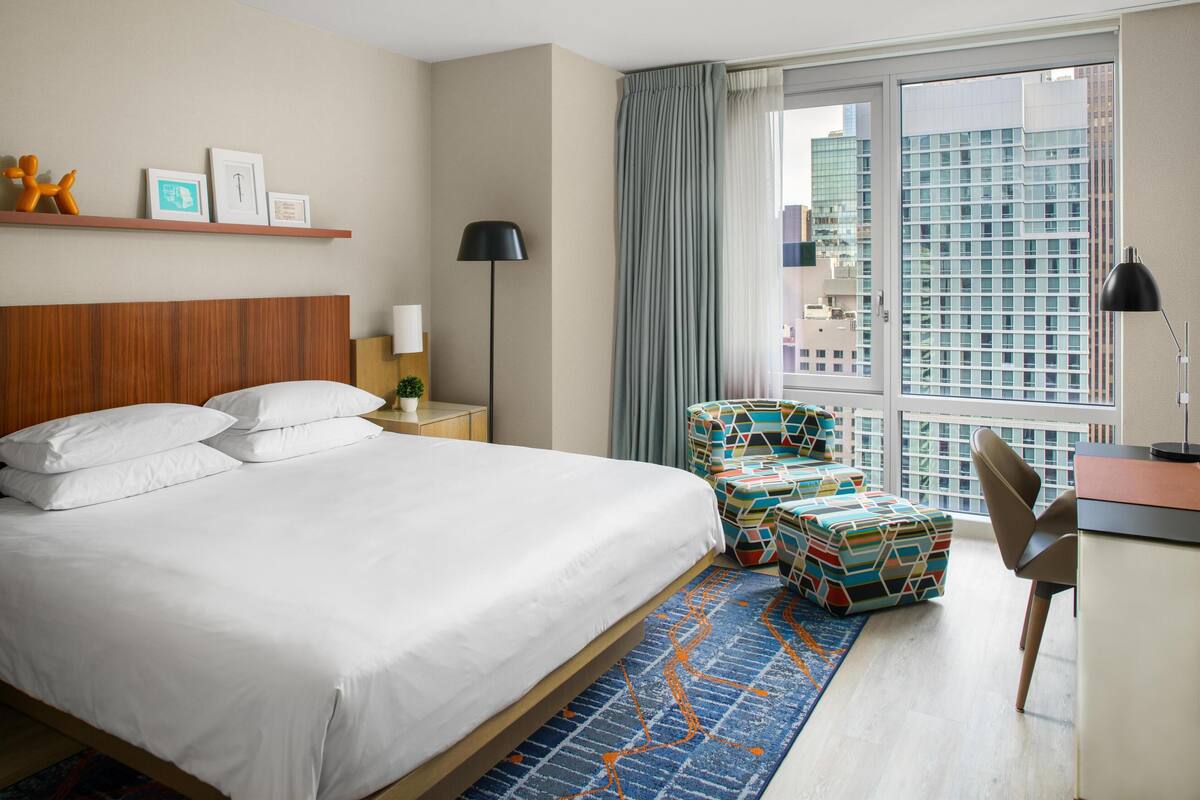 Quarto de hotel com uma cama grande, uma escrivaninha com cadeira, uma janela com vista para edifícios e um tapete estampado.