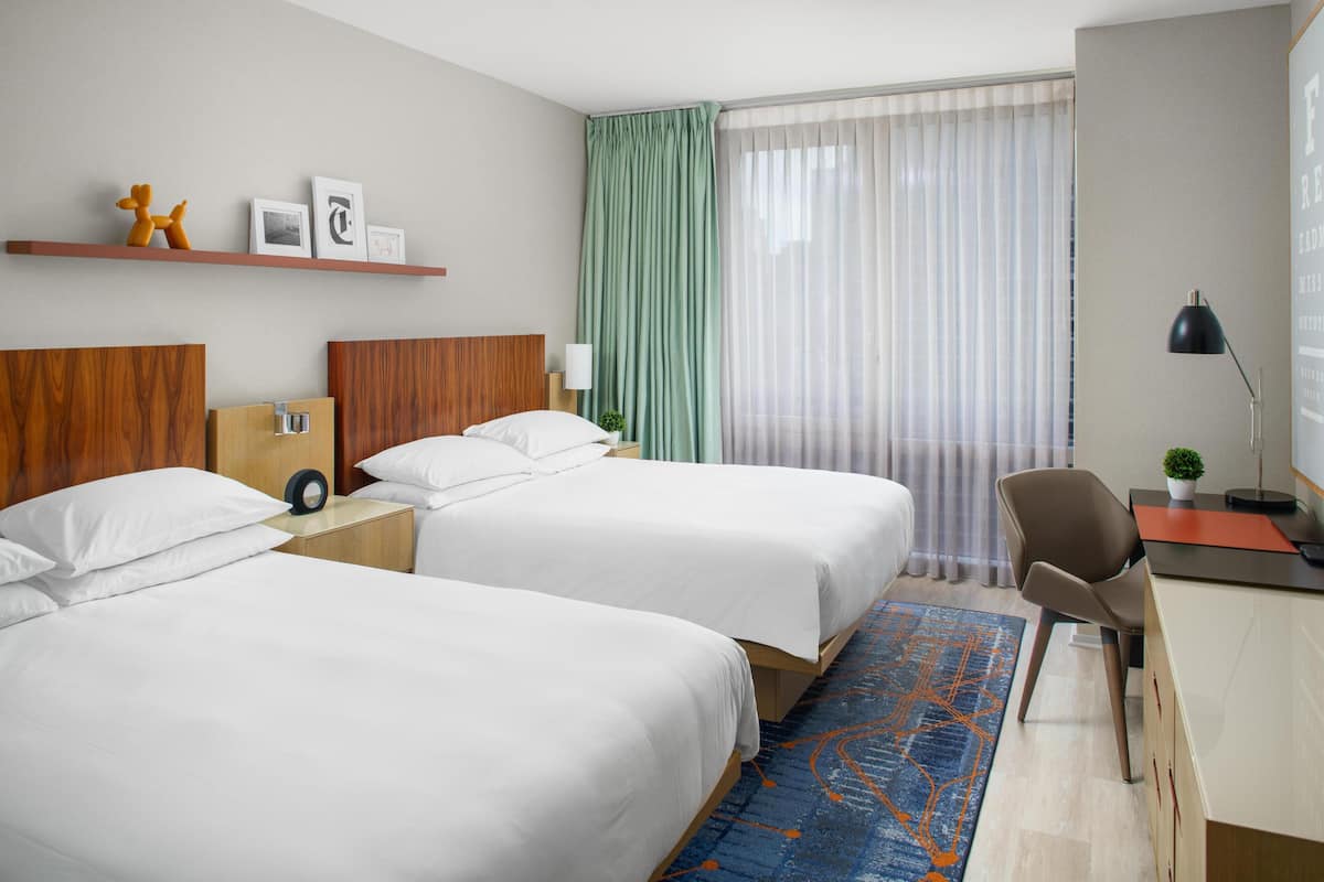 Quarto de hotel com duas camas, uma escrivaninha, uma cadeira, um abajur, uma planta e uma janela com cortinas.