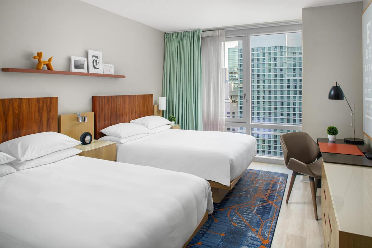 Quarto de hotel com duas camas, uma escrivaninha, uma cadeira, uma mesa de cabeceira, uma luminária, uma janela com vista para um edifício e um tapete azul estampado.