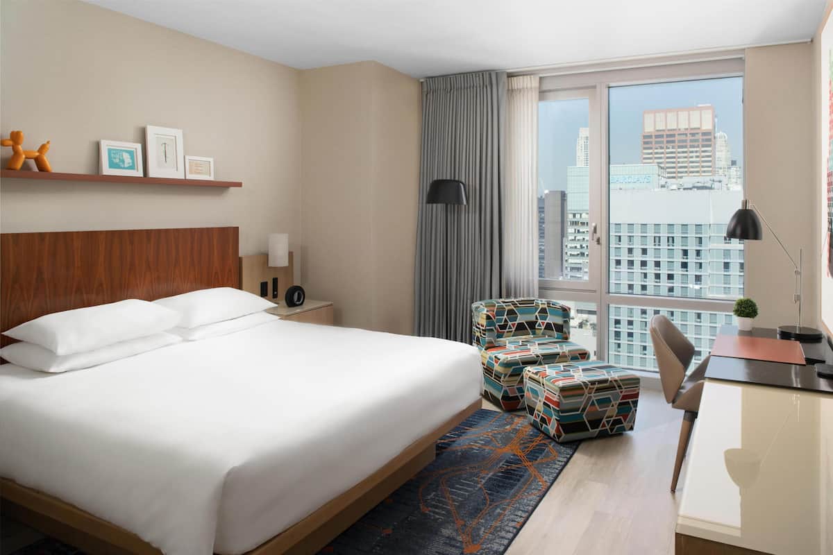 Quarto de hotel com uma cama grande, uma escrivaninha, uma cadeira, uma mesa pequena, um abajur, uma janela com vista para a cidade e uma estante com fotos e um brinquedo.