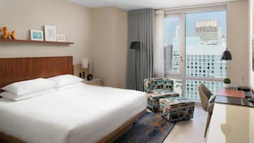 Habitación de hotel con una cama grande, un escritorio, una silla, una mesita, una lámpara, un ventanal con vistas a la ciudad y un estante con fotografías y un juguete.