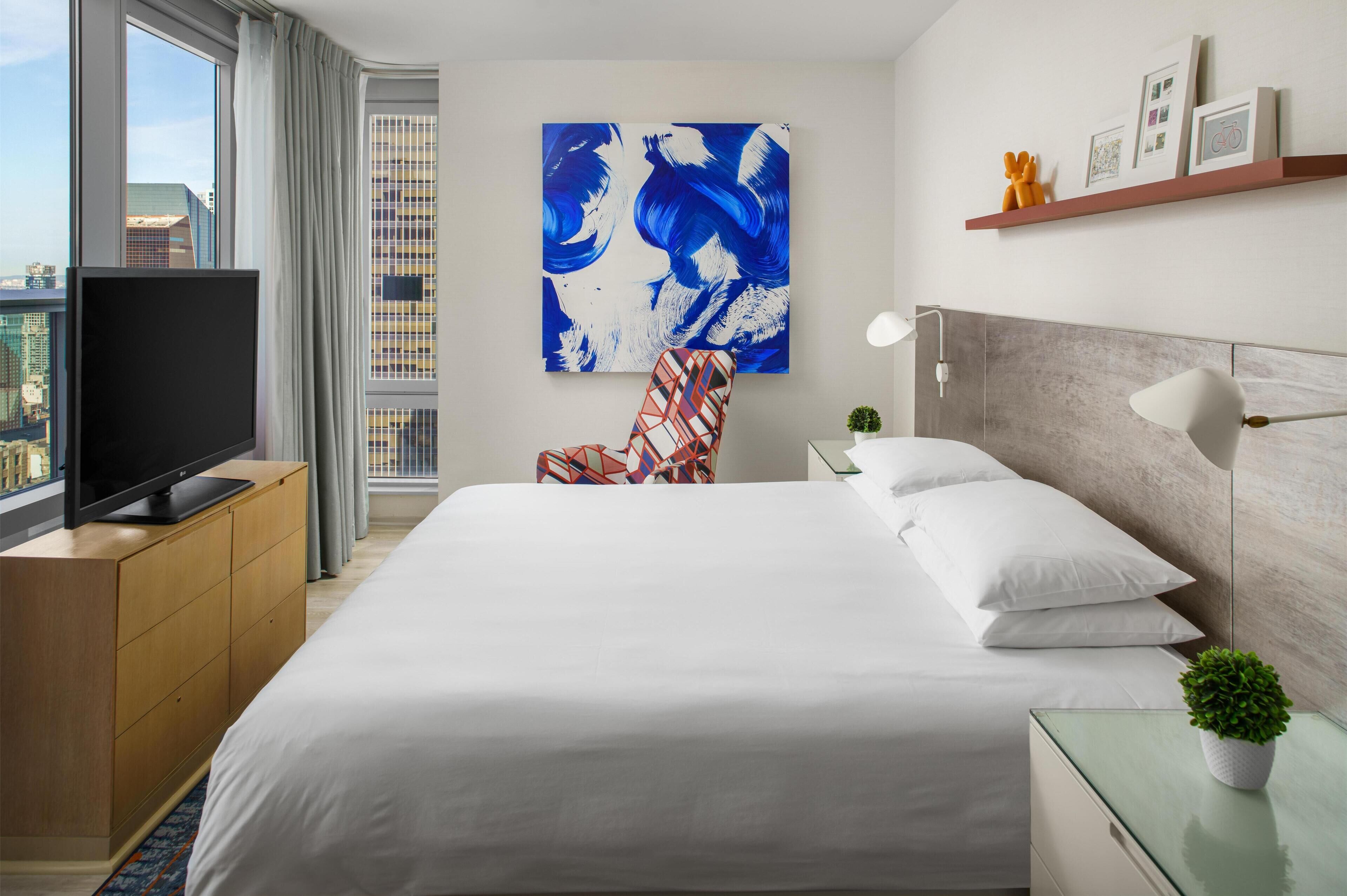 Une chambre d’hôtel avec un grand lit, un téléviseur posé sur un meuble en bois, une chaise à motifs géométriques, un tableau accroché au mur et une vue sur la ville par la fenêtre.