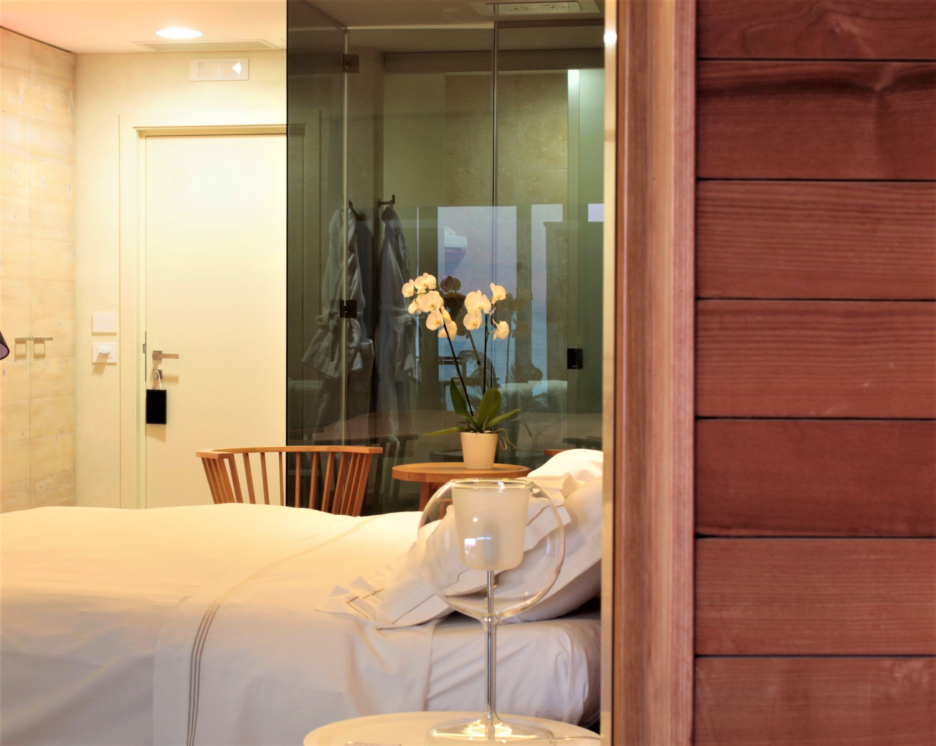 A hotel room with a bed, a wooden chair, a small table with a potted plant, and a glass partition.