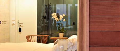 A hotel room with a bed, a wooden chair, a small table with a potted plant, and a glass partition.