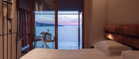 A bedroom with a bed, a nightstand, a lamp, and a view of the sea through a sliding door.