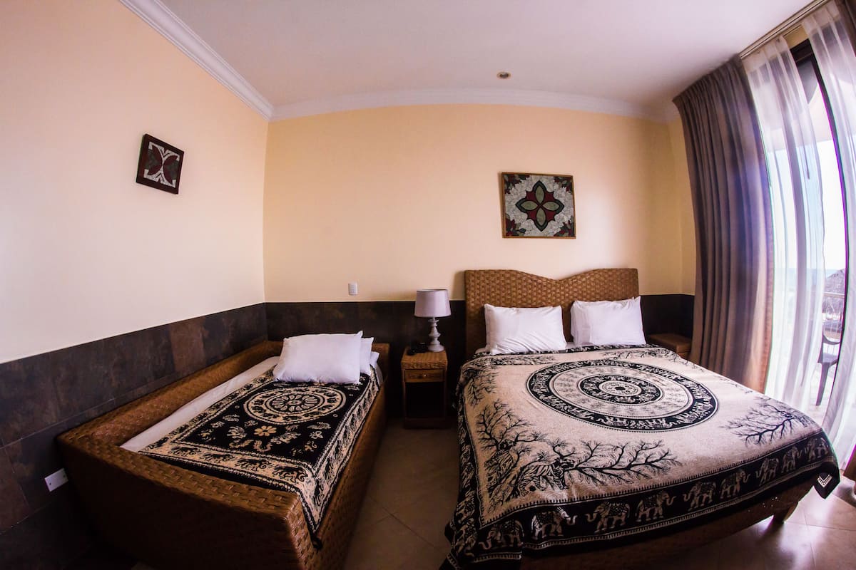 A hotel room with two single beds, a nightstand with a lamp, a decorative wall hanging, and a large window with curtains.