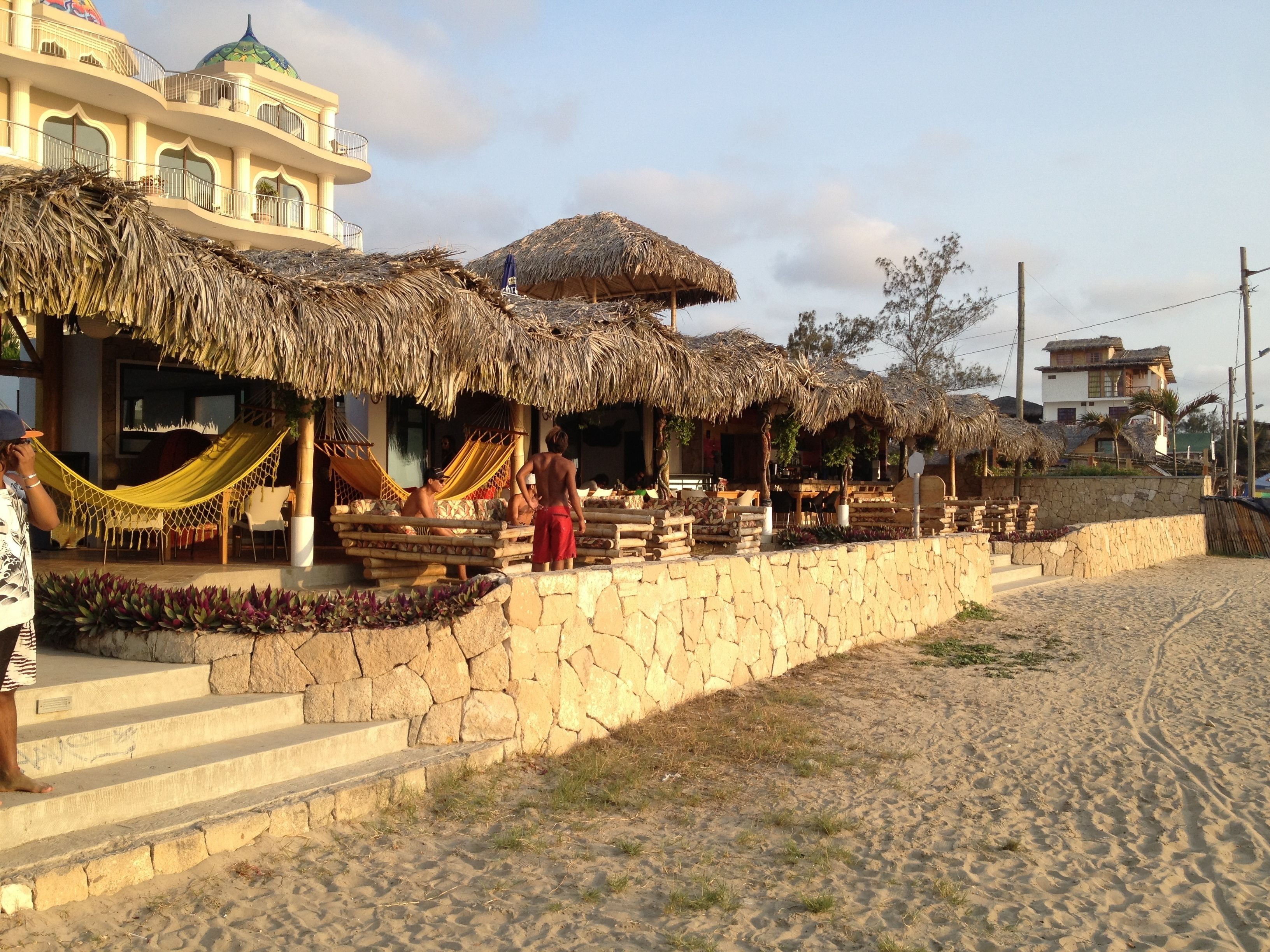 A beachfront property with thatched-roof structures, a stone wall, and a sandy beach.