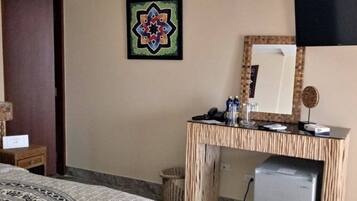 A hotel room with a bed, a desk with a mirror, a small refrigerator, and a decorative wall piece.