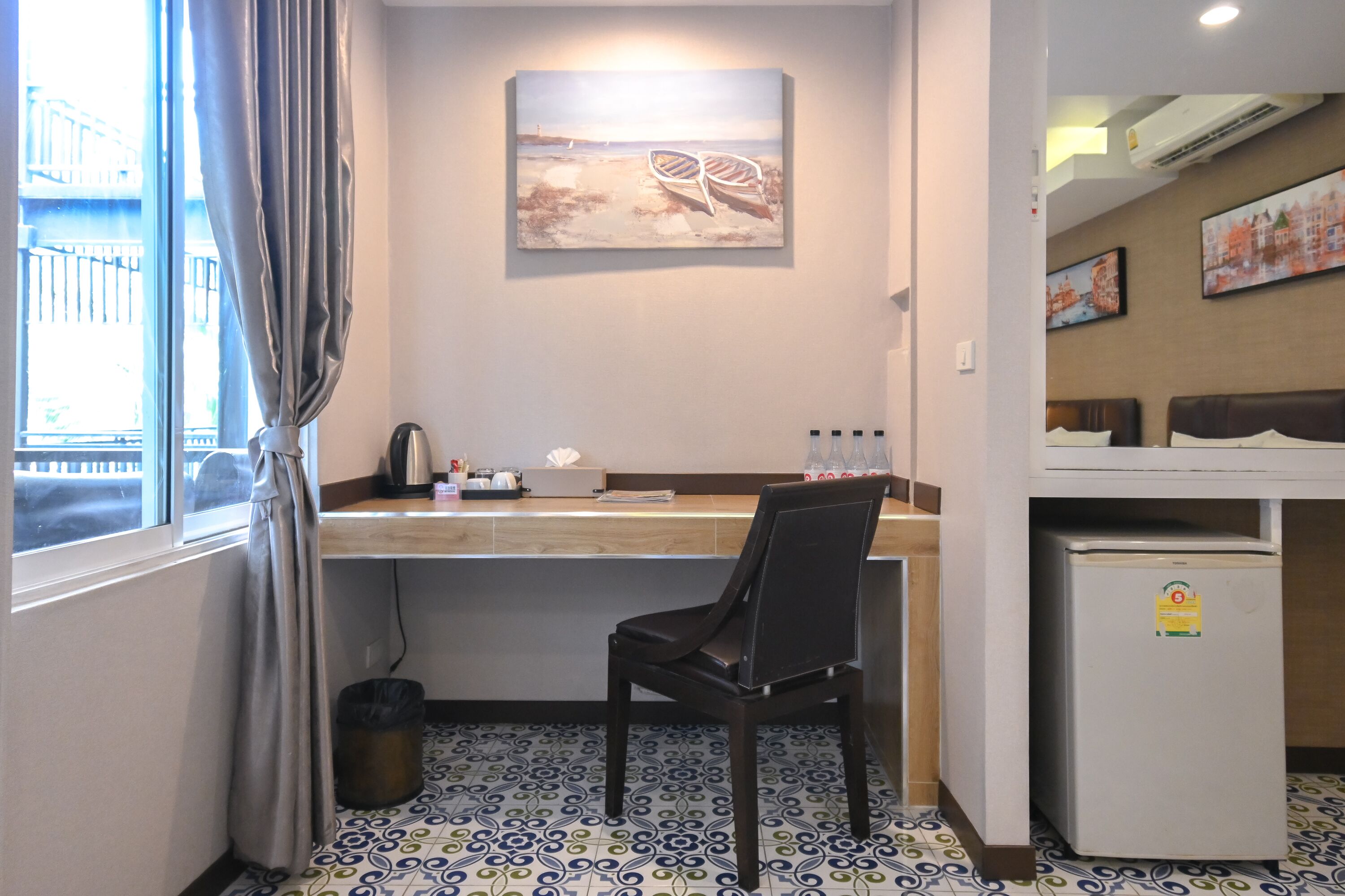 A hotel room with a desk, chair, and a small refrigerator. There is a window with curtains, a painting on the wall, and a tiled floor.