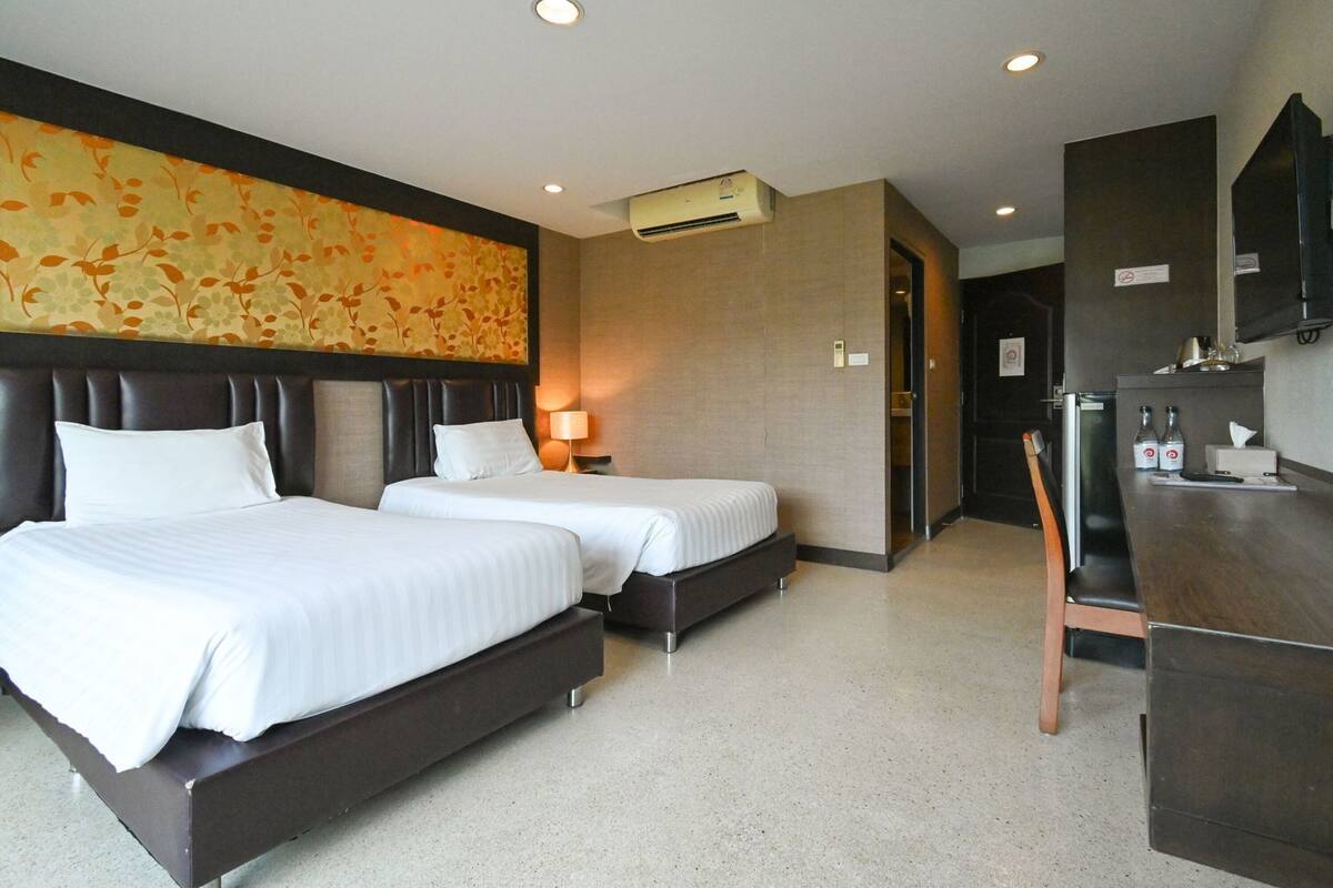 Ein Hotelzimmer mit zwei Betten, einem Schreibtisch, einem Stuhl, einer Klimaanlage und einer Wand mit einem Blumenmuster.