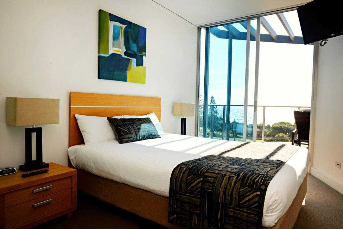 Habitación de hotel moderna con una cama grande, una mesita de noche con lámpara y vistas al mar a través de amplios ventanales.