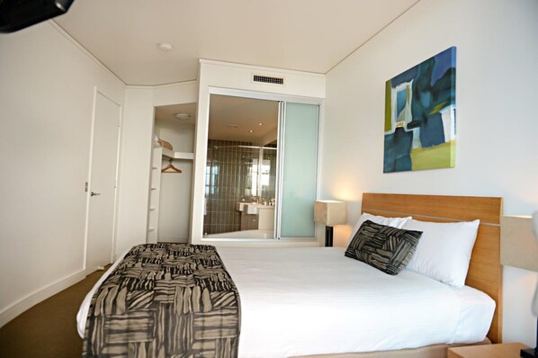Habitación de hotel con cama, mesita de noche, lámpara de pared, un cuadro y una puerta corredera que da al baño.