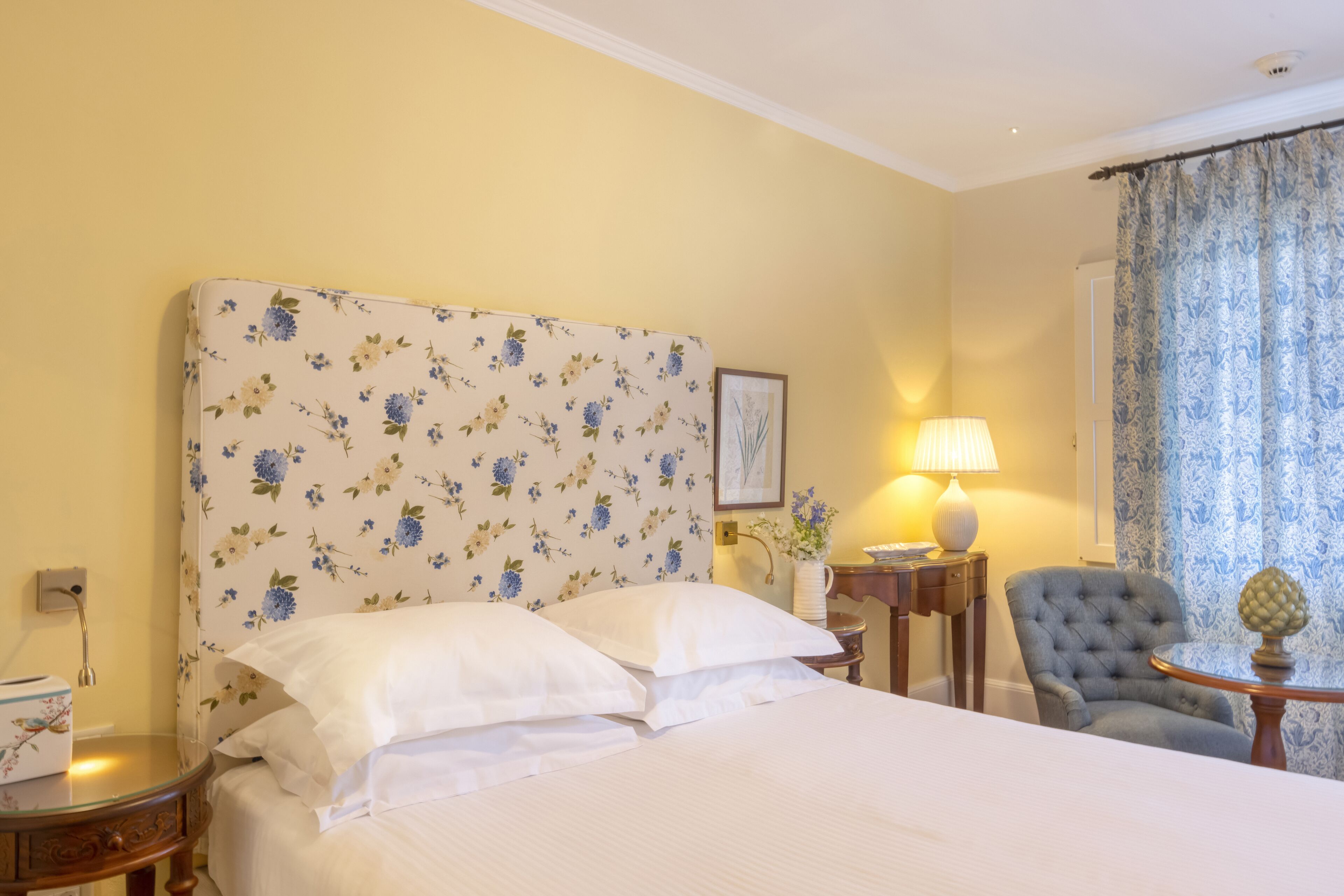 Una habitación de hotel con un cabecero floral, una cama con sábanas blancas, un sillón gris, una mesita de noche con una lámpara y una ventana con cortinas estampadas.