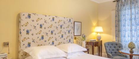 Una habitación de hotel con un cabecero floral, una cama con sábanas blancas, un sillón gris, una mesita de noche con una lámpara y una ventana con cortinas estampadas.