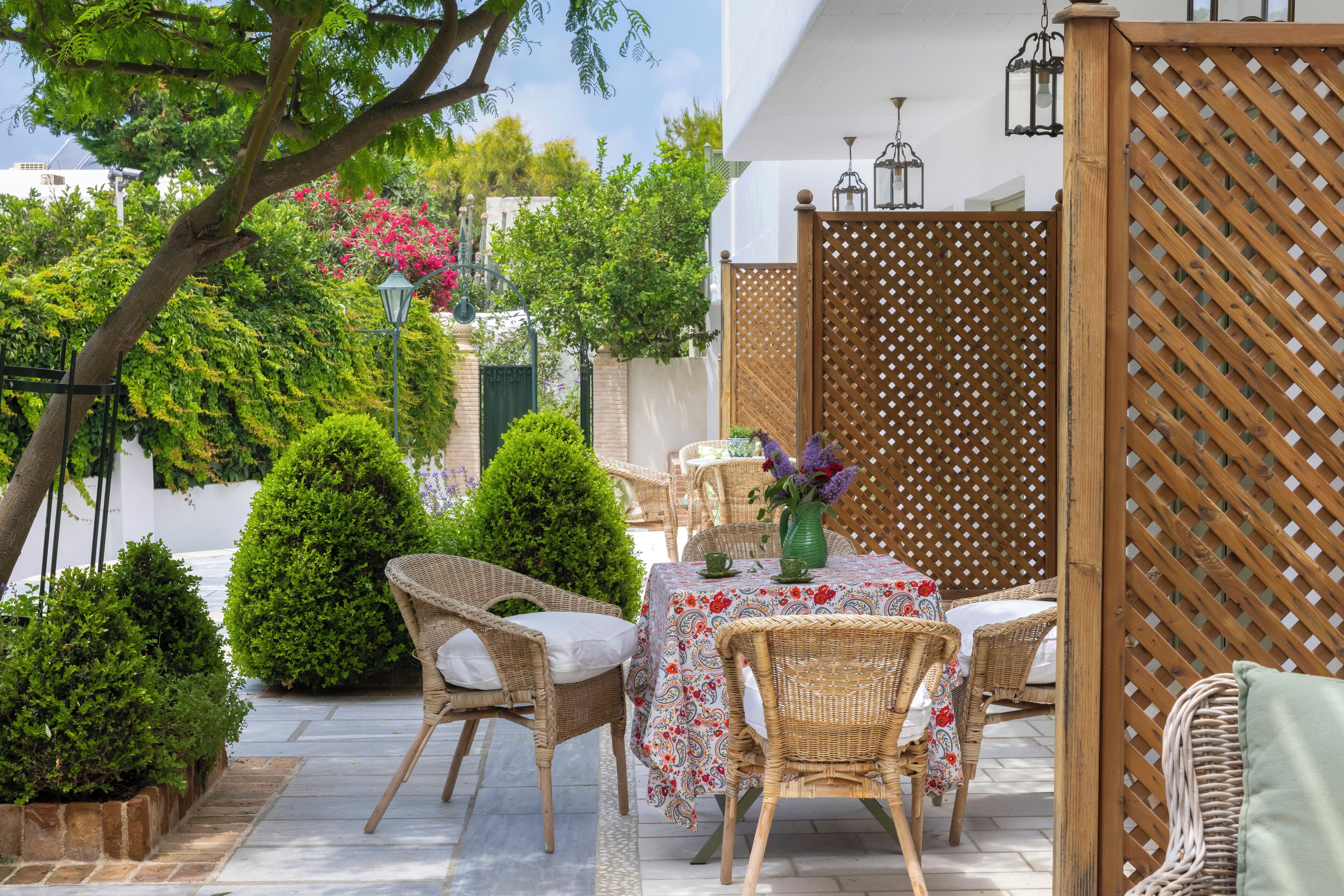 Een terras met rieten meubels, een tafel met een bloemrijk tafelkleed en een houten schutting.