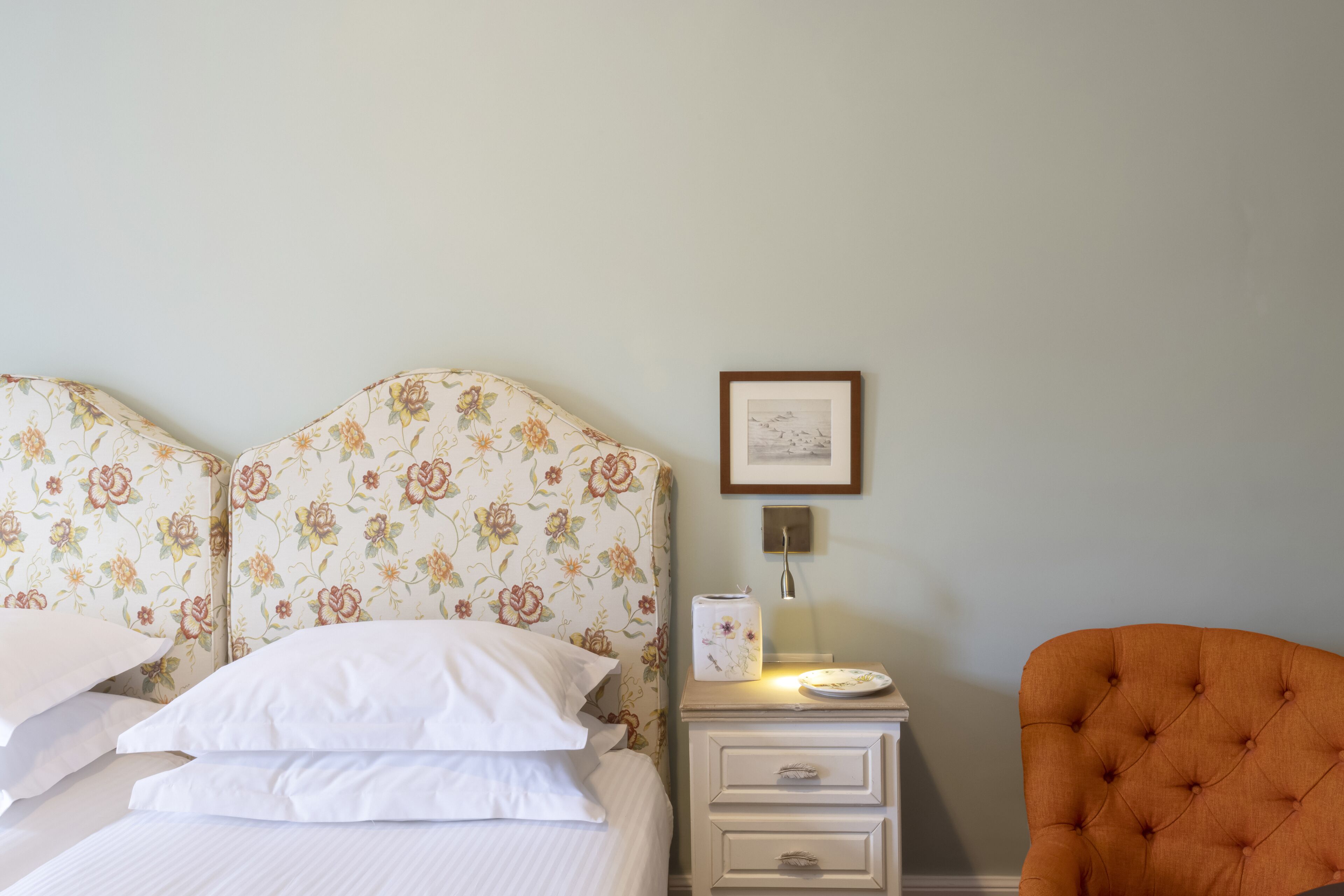 A neatly made bed with floral headboard, a bedside table with a lamp, and an orange chair.