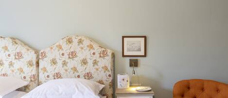 A neatly made bed with floral headboard, a bedside table with a lamp, and an orange chair.