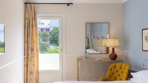 A hotel room with a bed, a yellow armchair, a bedside table with a lamp, a mirror, and a view of greenery outside. - Kouneni Apartments (Mykonos)